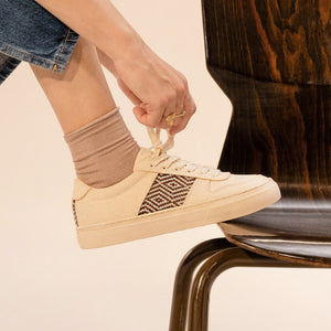 NGO SHOES Handgemachte beige Sneaker mit braunem Ethno-Muster, getragen von einer Person, die die Schnürsenkel bindet.
Nachhaltige und vegane Fair-Trade-Schuhe auf einem stilvollen Holzstuhl.