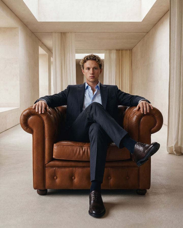 Man in a suit sitting on a brown leather armchair in a bright room with high ceilings.