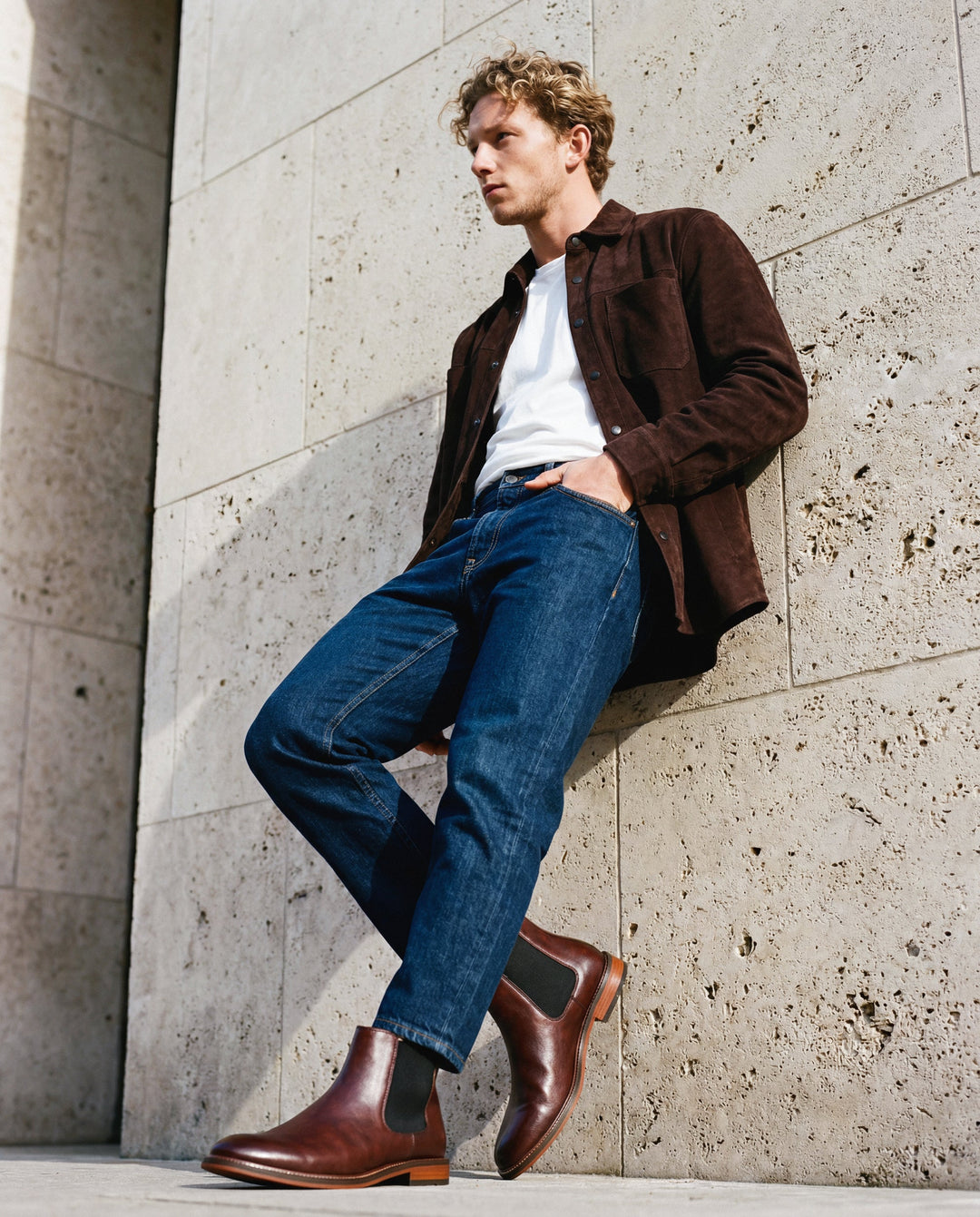 Man wearing a brown jacket, white shirt, blue jeans, and brown vegan boots from nae vegan shoes leaning against a stone wall.