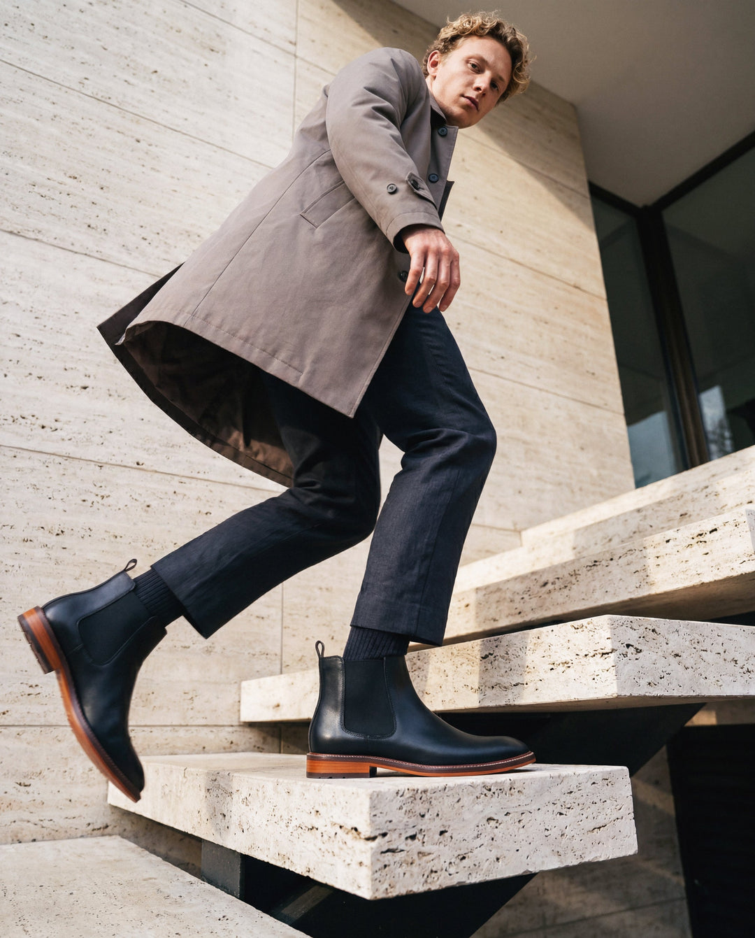 Man in a gray coat and black boots sitting on concrete steps.