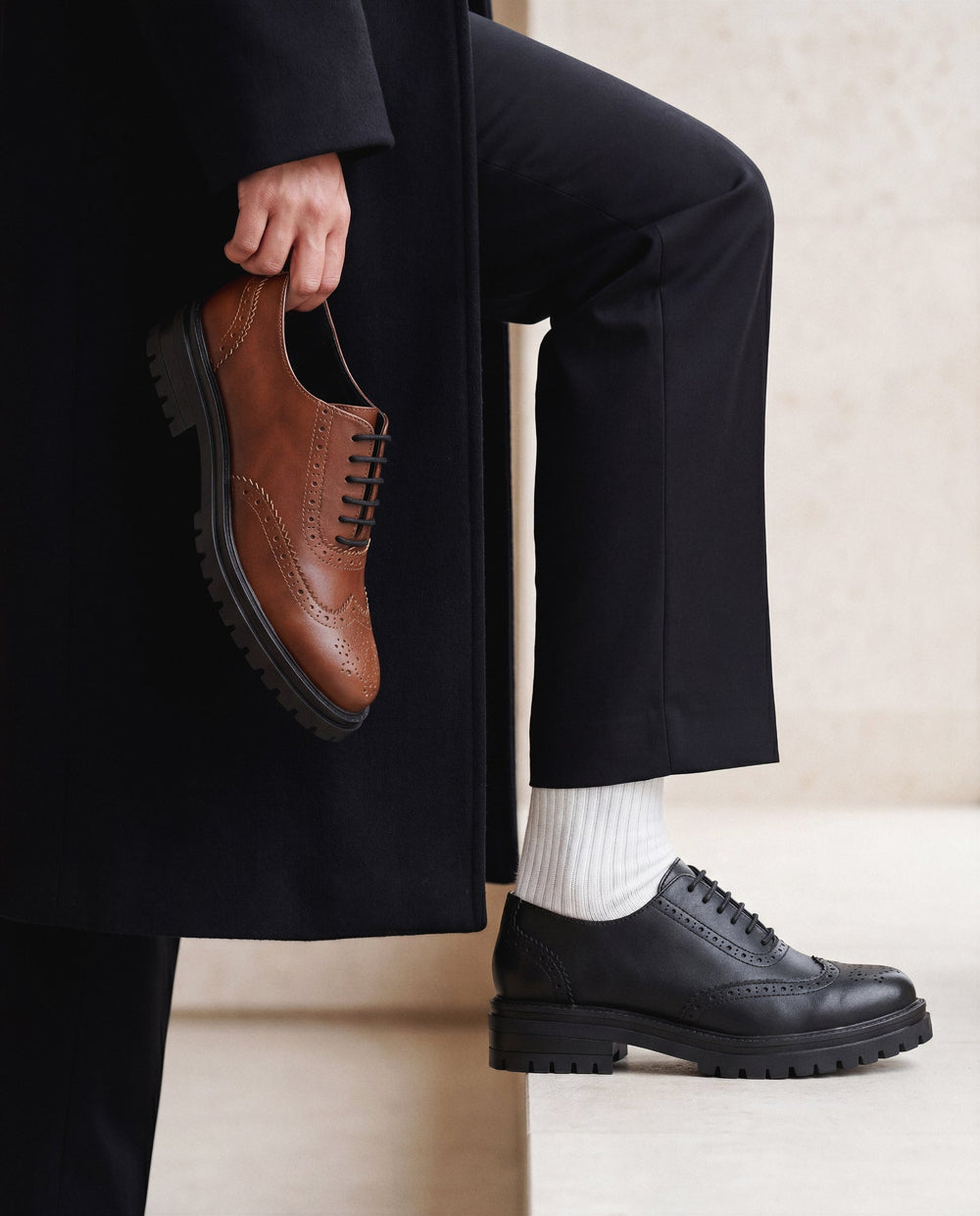 Person wearing black shoes and holding a brown shoe, with a neutral background