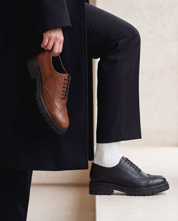 Person wearing black shoes and holding a brown shoe, with a neutral background